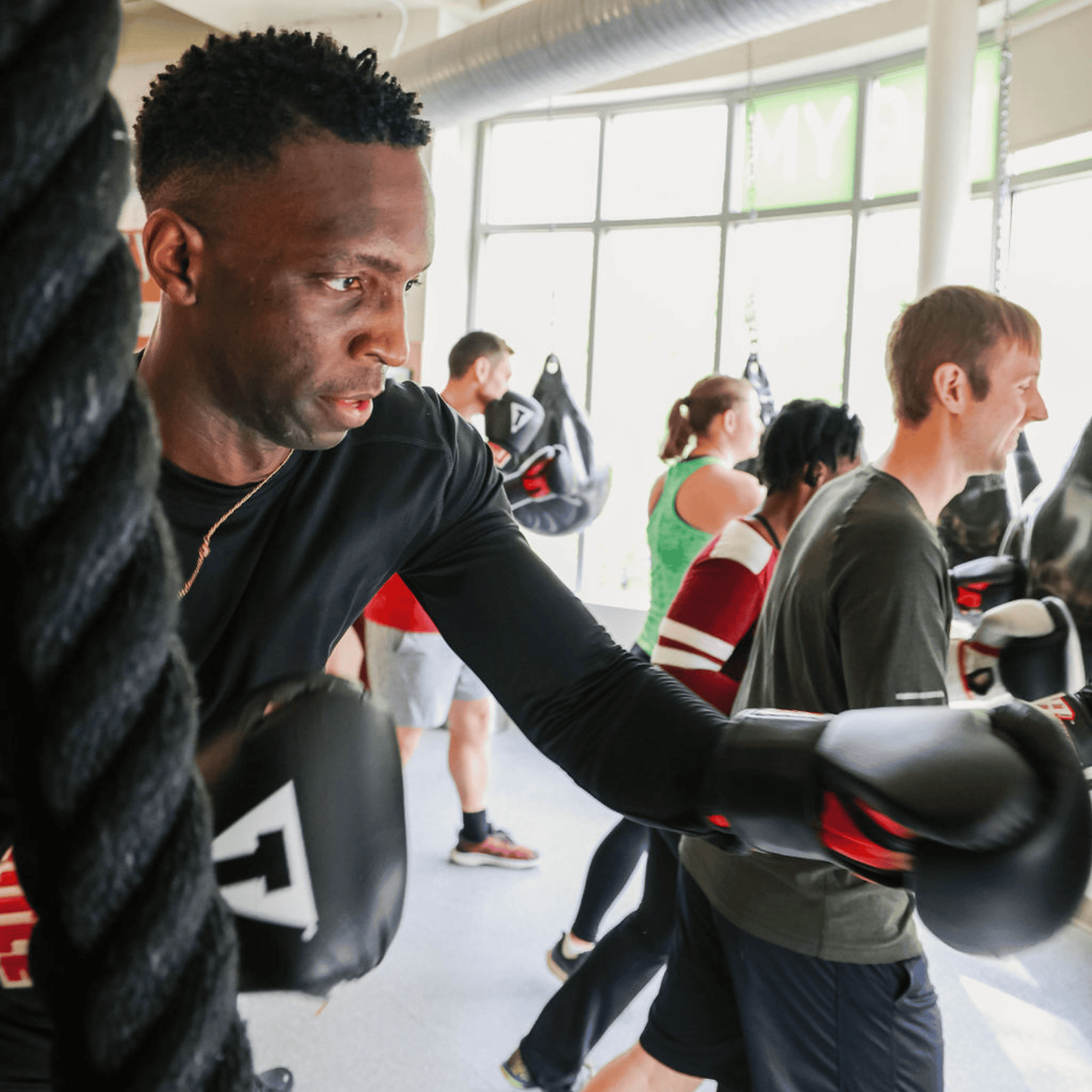 A member wearing boxing gloves works a battle rope during a high-intensity boxing fitness class in MINT's natural light-filled studio in Adams Morgan, Washington DC, with other members sparring in the background.