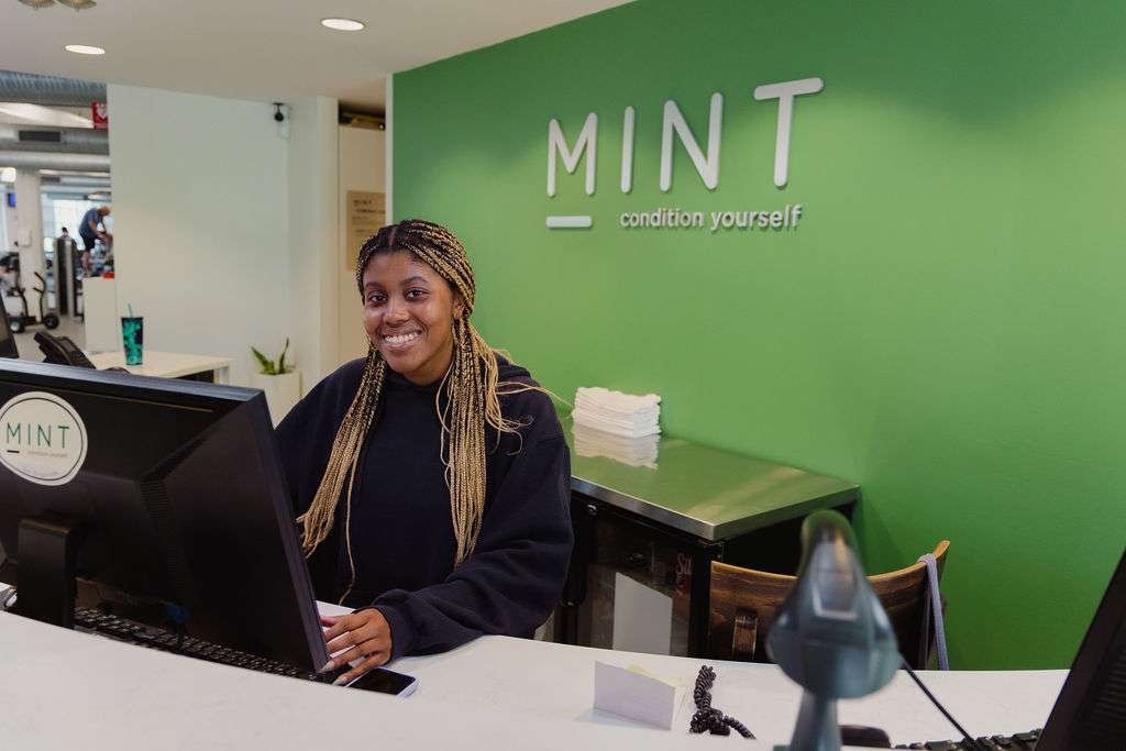 A friendly staff member smiles behind the front desk at MINT gym in Adams Morgan DC, with the signature green MINT Condition Yourself wall and gym floor visible in the background